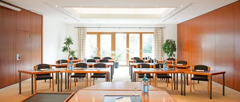 A conference room with wooden tables and chairs, large windows, and potted plants.