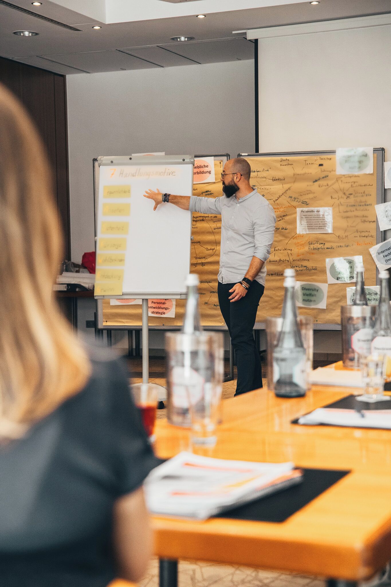 A man presenting in a meeting room with a whiteboard, papers, and a table with documents and bottles.