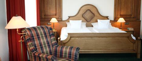 Una habitación de hotel con una cama de madera, dos lámparas de noche, un sillón escocés y un reposapiés a juego.