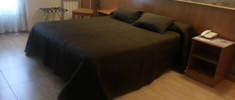 A hotel room with a large bed, a wooden nightstand with a telephone, a chair, and a radiator.