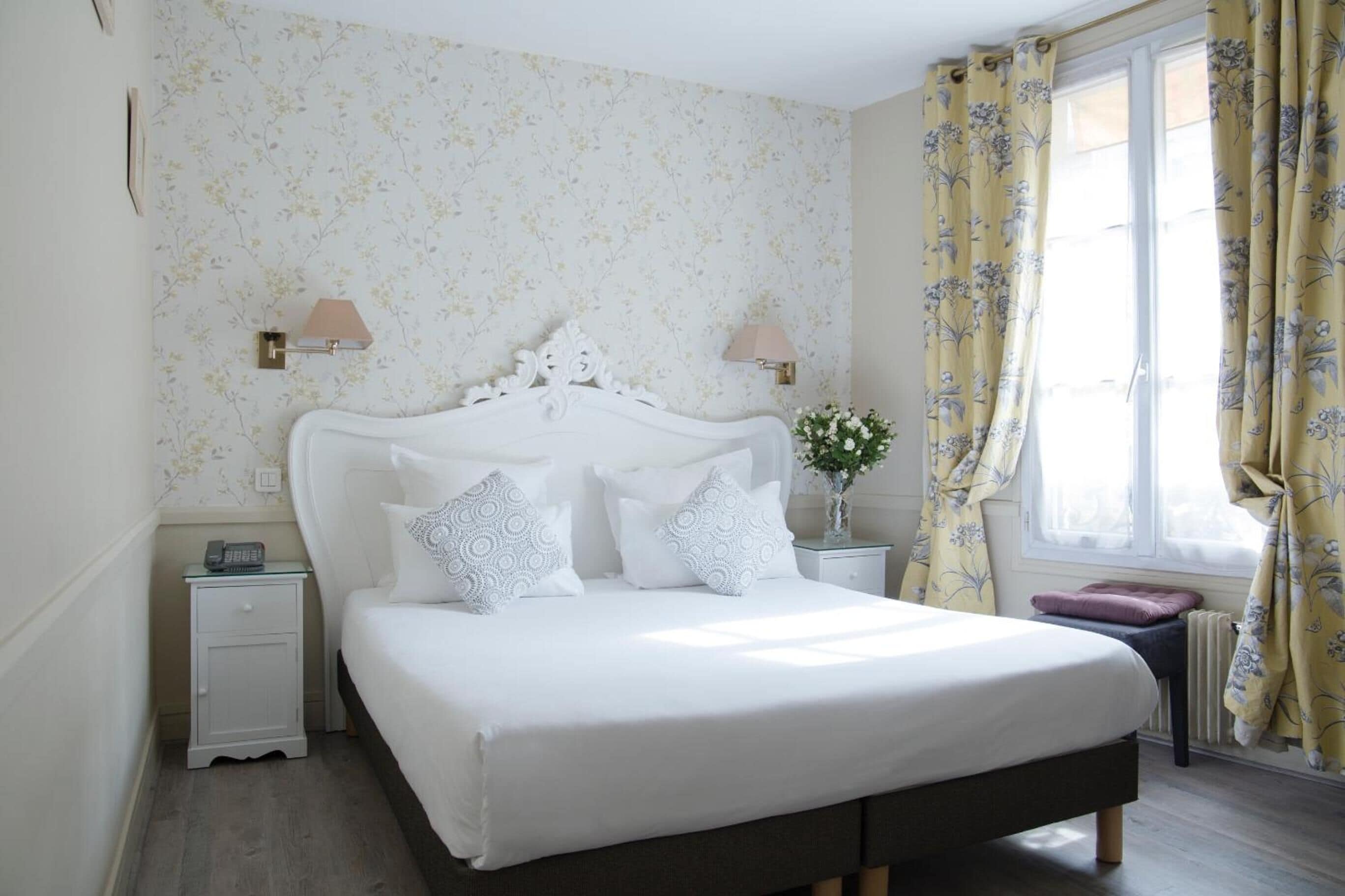Ein Schlafzimmer mit einem weißen Bett, zwei Nachttischlampen, einem Nachttisch, einem kleinen Tisch mit einer Vase und einem Fenster mit Vorhängen.