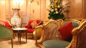 Una habitación con muebles ornamentados, incluyendo un sillón verde, un sofá rojo y dorado, y un sillón a juego con un cojín verde. Hay un jarrón floral sobre una mesita.