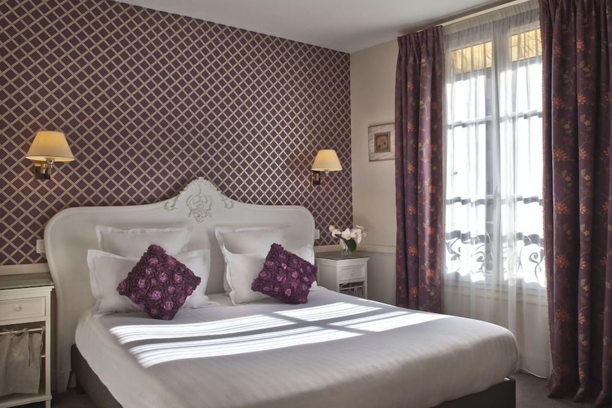 Un lit bien fait, avec du linge de lit blanc et des coussins à motifs floraux violets, une table de chevet avec une lampe et une fenêtre avec des rideaux.
