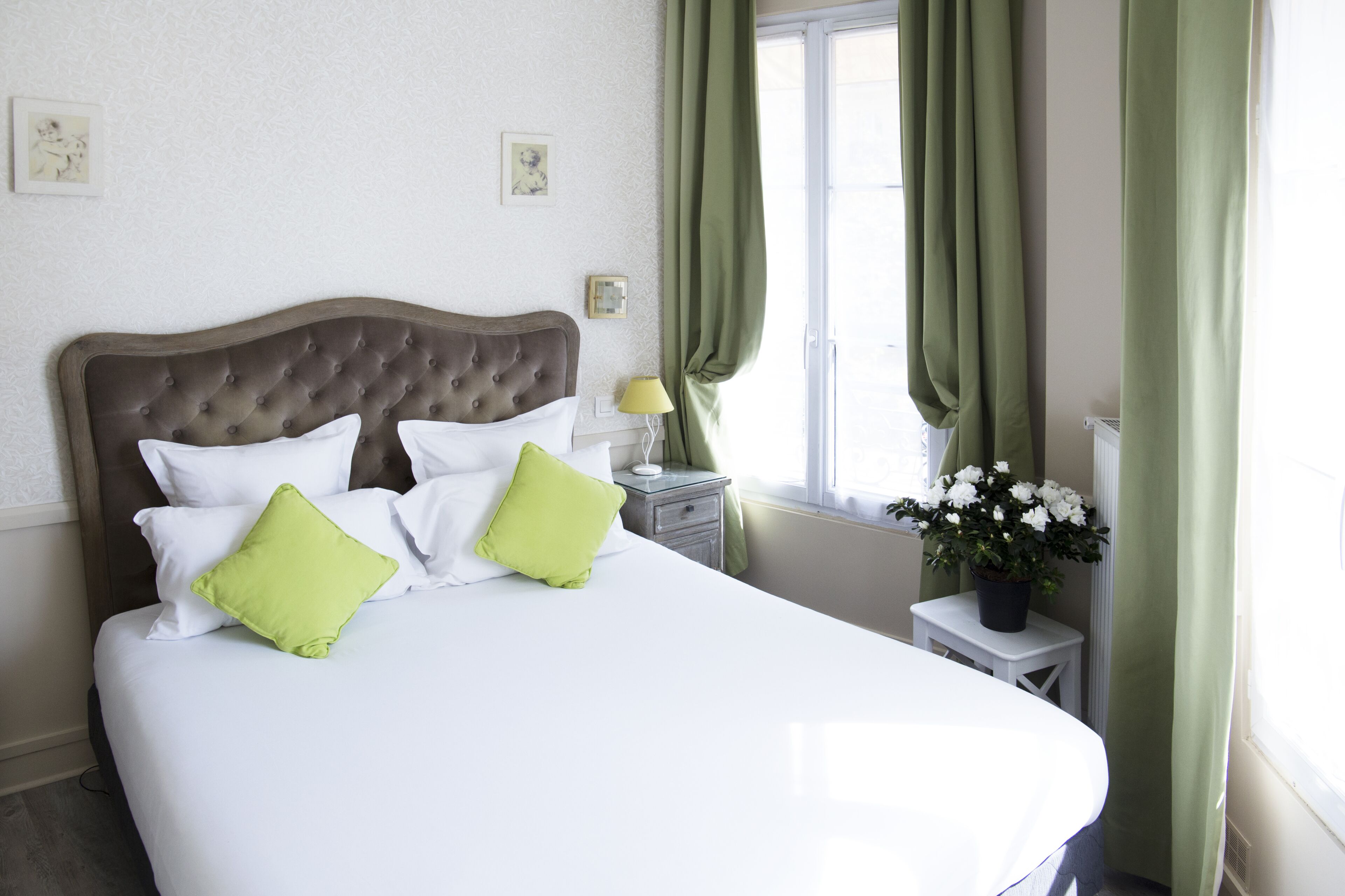 Ein ordentlich bezogenes Bett mit weißen und grünen Kissen, ein Nachttisch mit Lampe und ein Fenster mit grünen Vorhängen.
