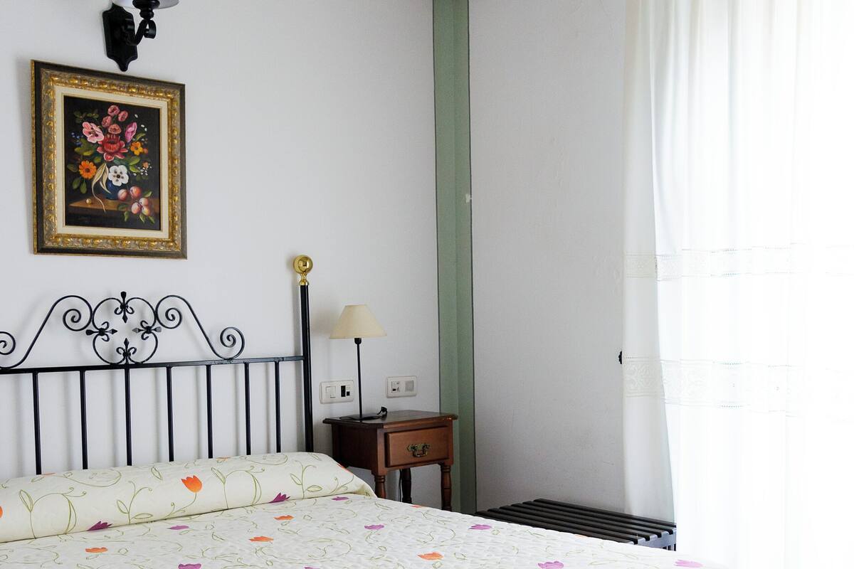 A bedroom with a bed, a nightstand, a lamp, a framed picture on the wall, and a window with curtains.