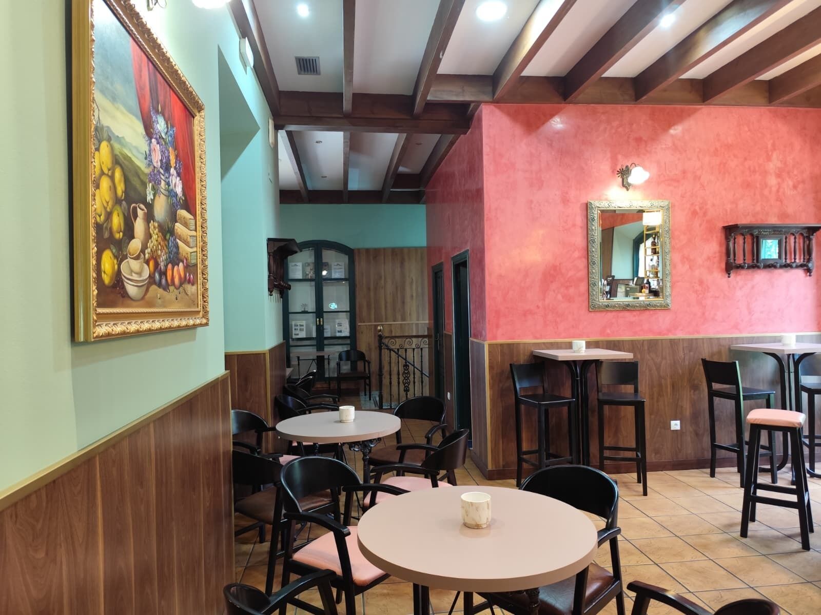 Interior de restaurante con mesas redondas, sillas negras, un cuadro, un espejo y techo de madera.
