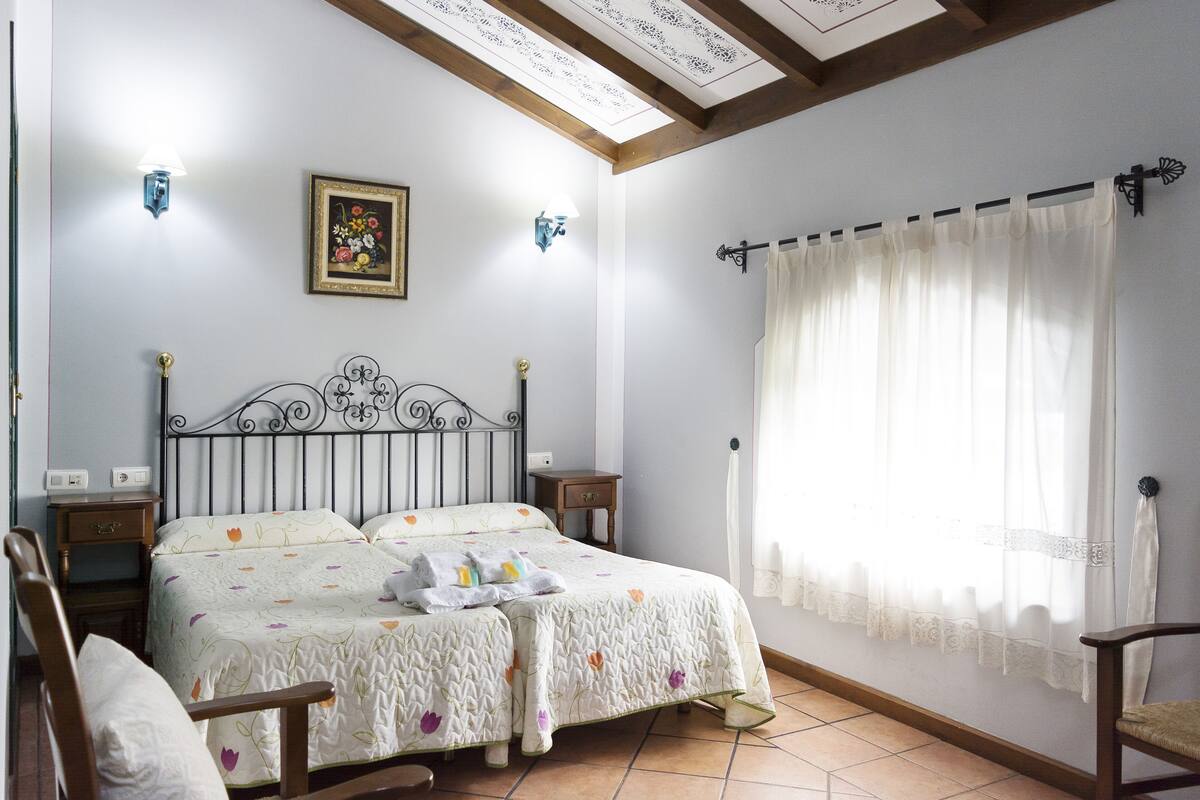 A bedroom with a bed, two bedside tables, a nightstand, a chair, a window with curtains, and a wall-mounted picture.