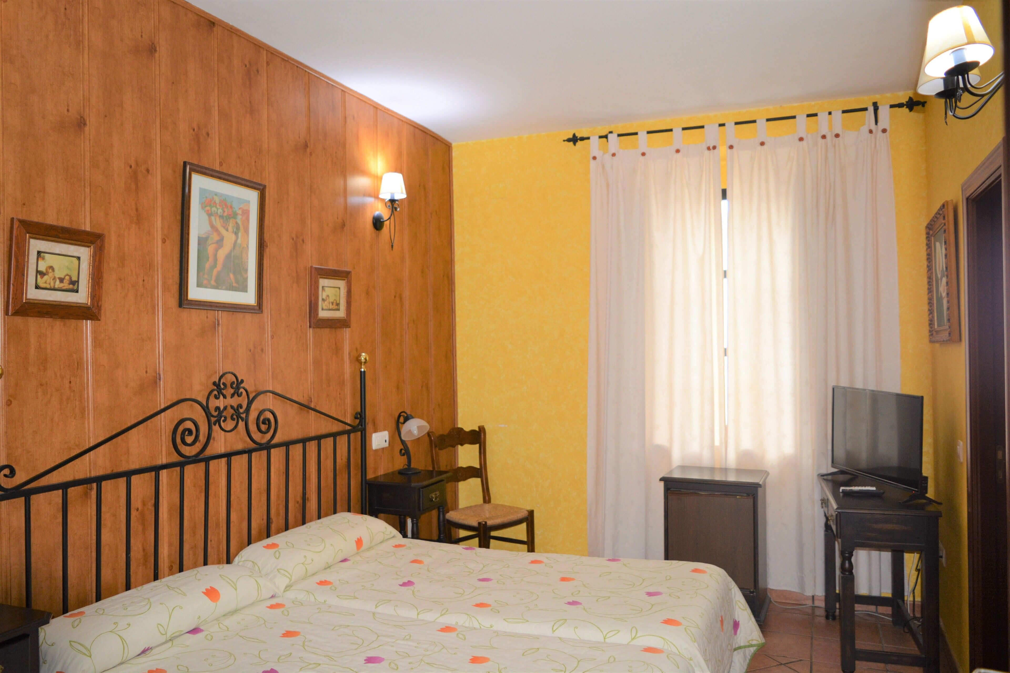 Una habitación de hotel con cabecera de madera, una cama con ropa de cama floral, una silla, una mesita con lámpara, un televisor sobre un mueble bajo y una ventana con cortinas.