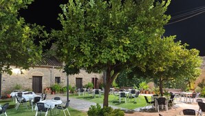 Un área de comedor al aire libre con suelo de piedra, mesas y sillas, rodeada de follaje y un edificio de piedra.