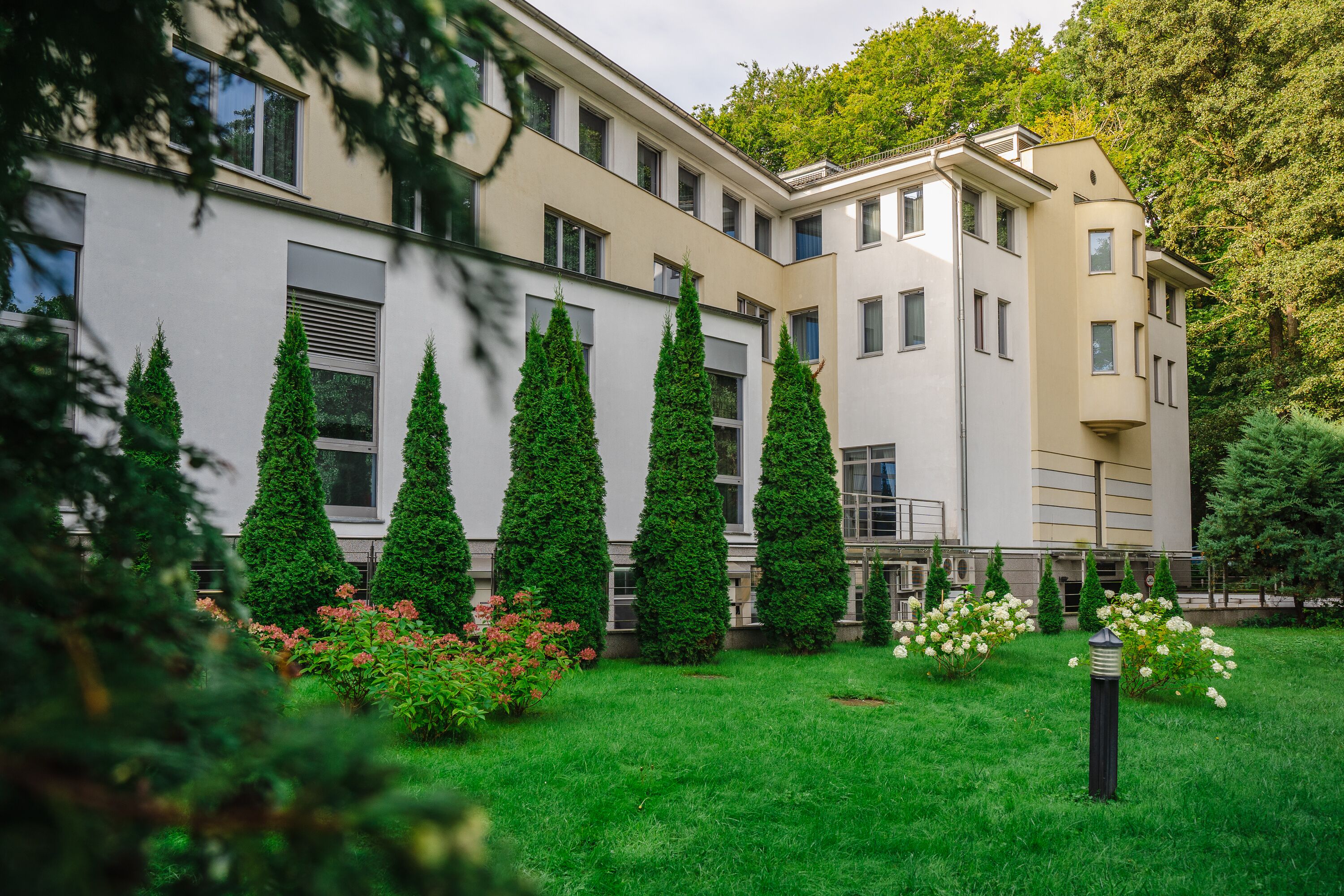 A modern building with a symmetrical facade, multiple windows, and a well-maintained garden with green hedges and flowering plants.