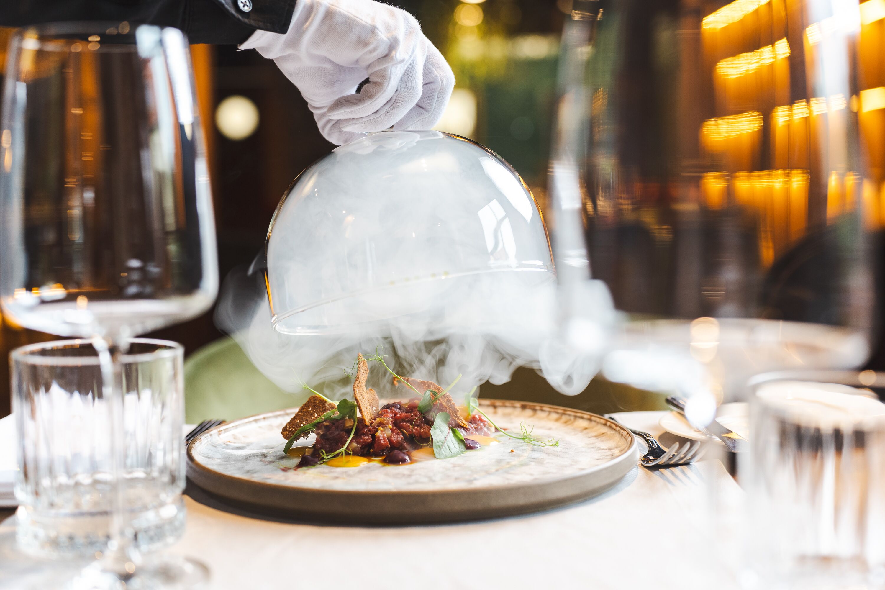 A waiter pouring a hot beverage over a dish, steam rising from the cup.