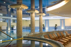 A large indoor pool area with multiple columns, lounge chairs, and a glass barrier.