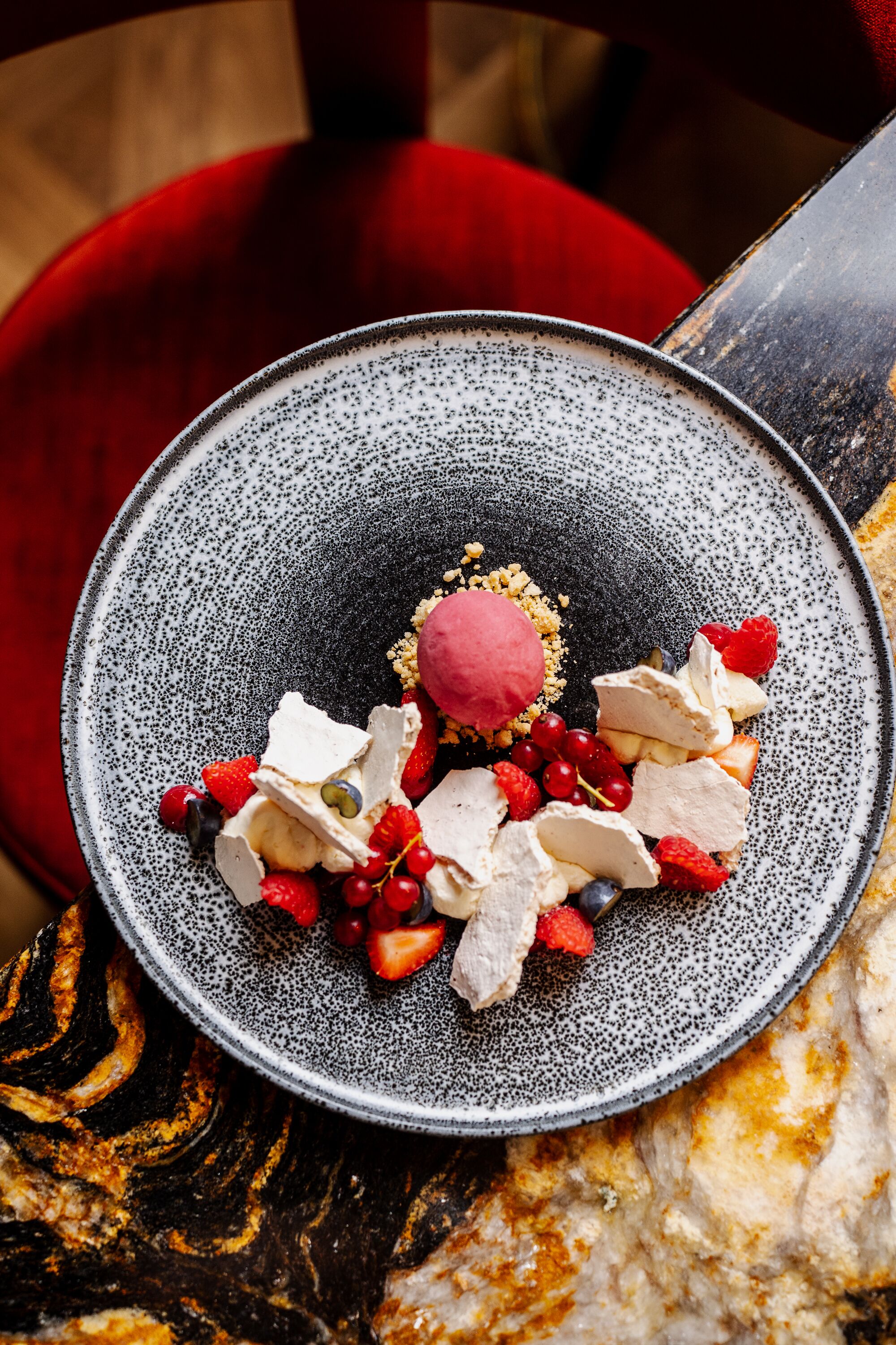 Un plato de postre con diversas frutas y una bola de helado sobre una fuente de cerámica con motas.