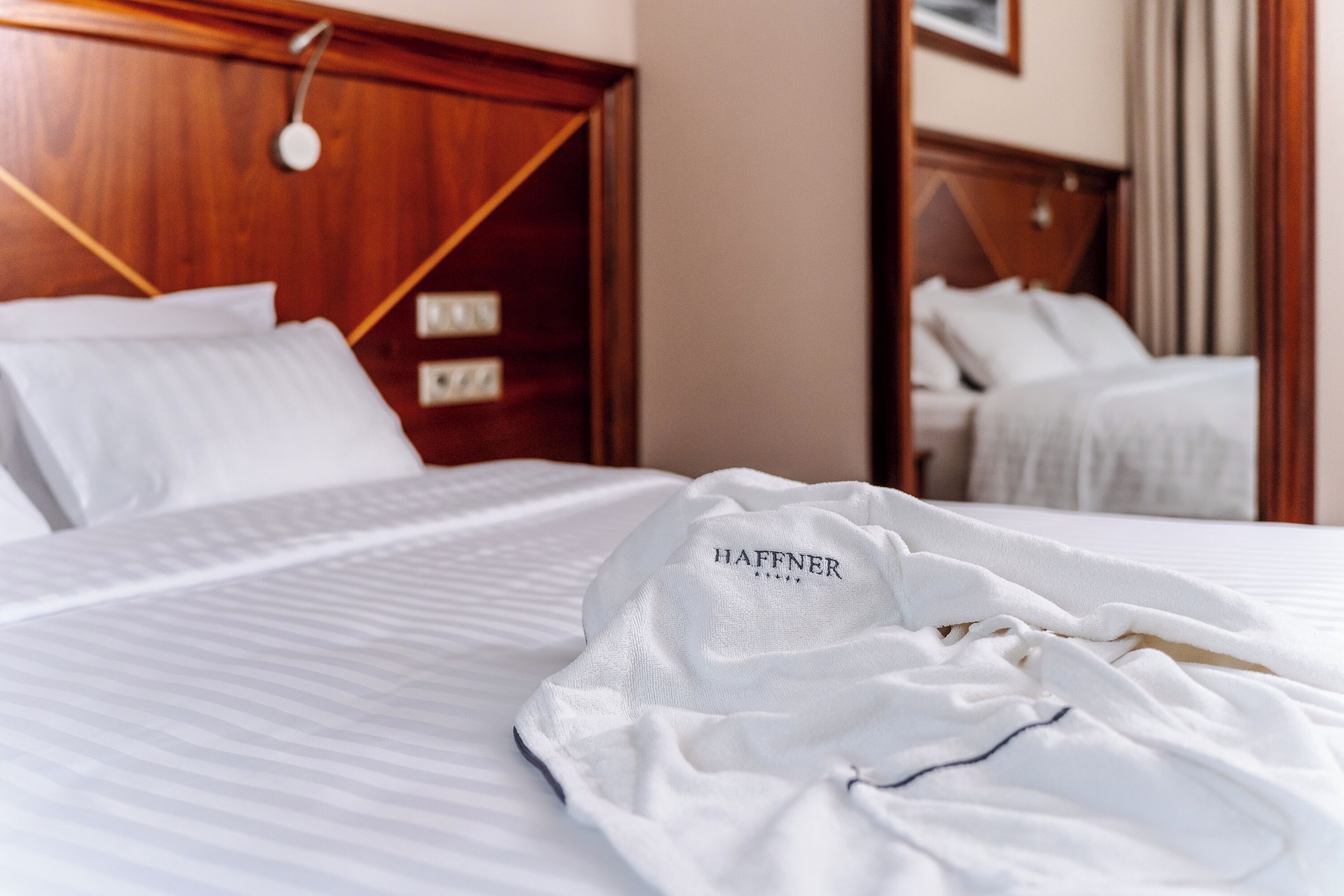 A hotel room with a neatly made bed, a wooden headboard, and a white robe with 'HAFFNER' embroidered on it.