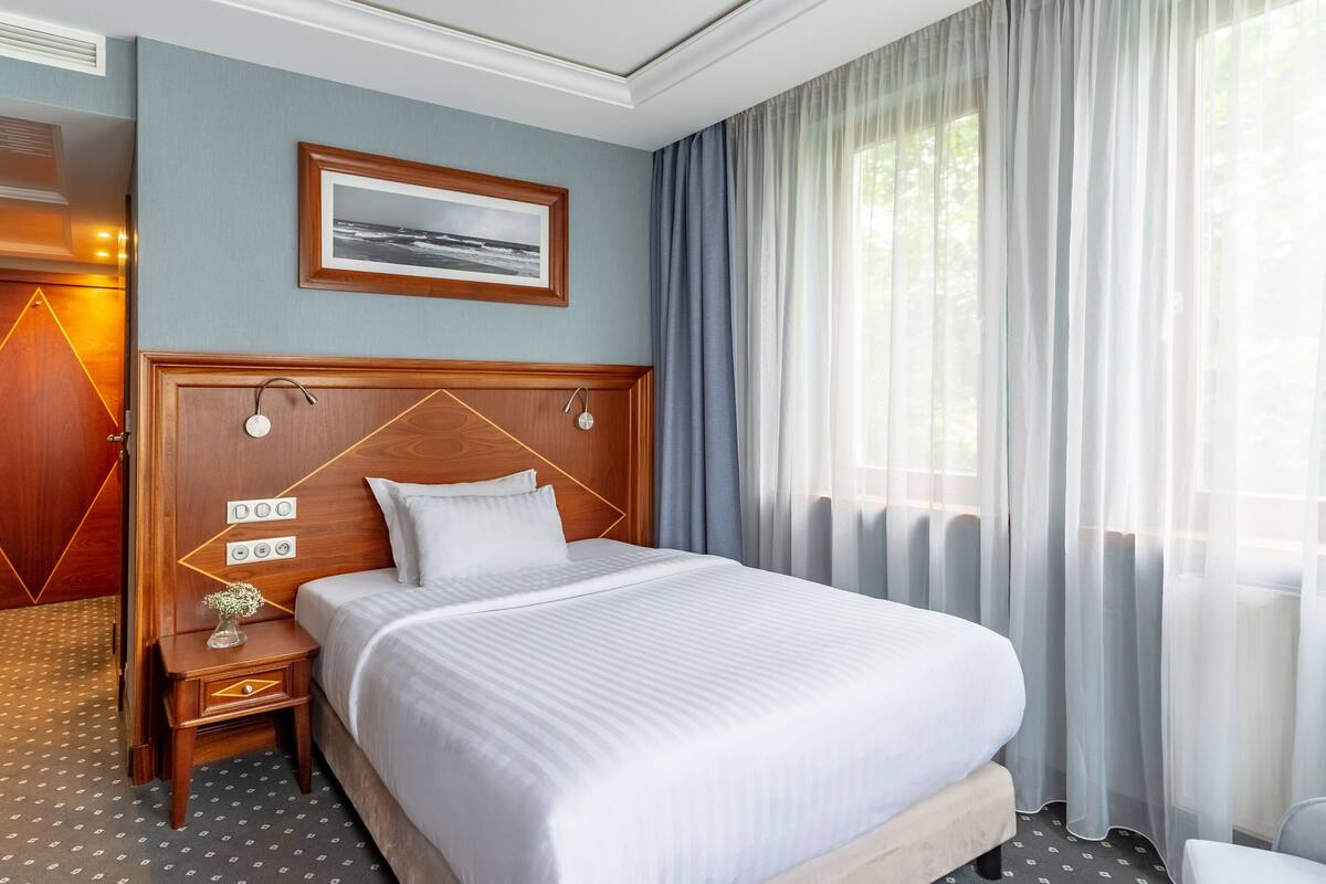 A hotel room with a large bed, a wooden headboard, a bedside table with a vase, and a window with sheer curtains.