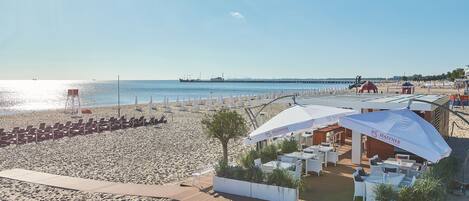 Un restaurant a la vora de la platja amb terrassa, vistes al mar i un moll al fons.