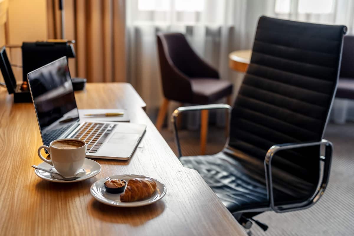 A wooden table with a laptop, a cup of coffee, and a plate with a croissant.