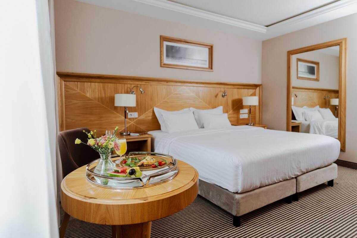 A hotel room with a bed, bedside tables, a round wooden table with a tray of food, and a mirror on the wall.