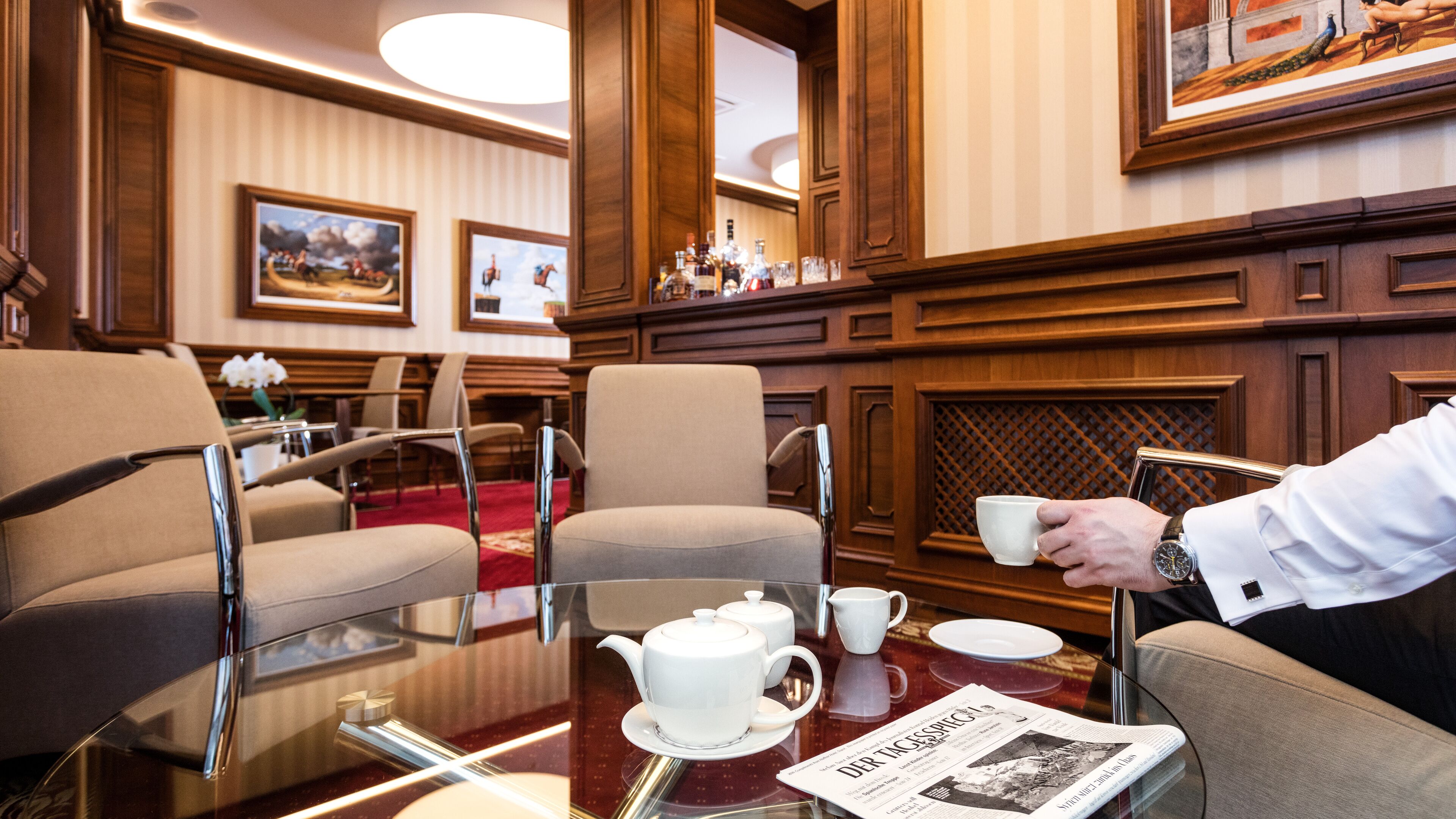 A hotel room with a glass table, a white teapot, a newspaper, and a framed painting on the wall.