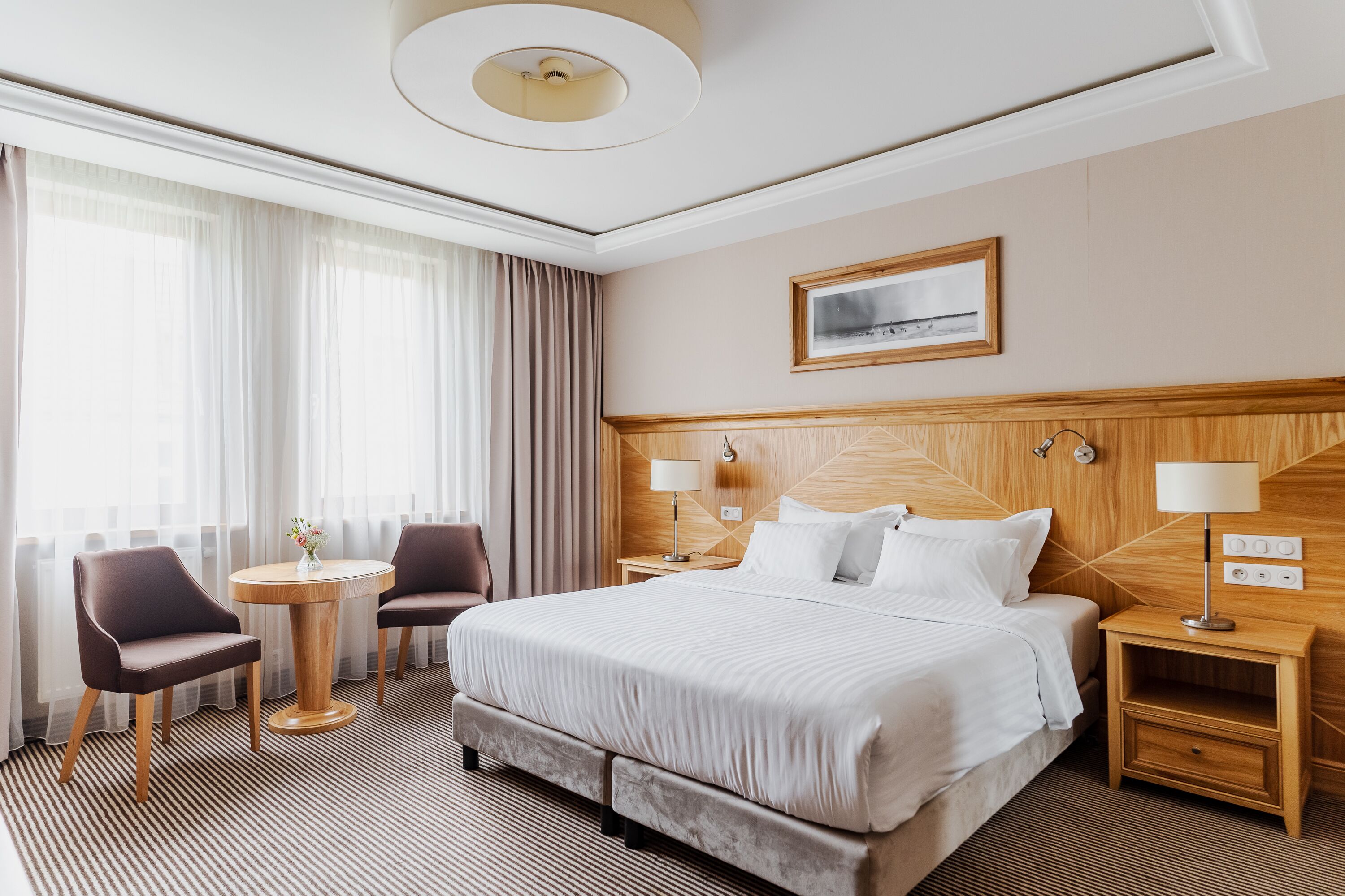 A hotel room with a large bed, two bedside tables, a round table with chairs, and a large window with curtains.