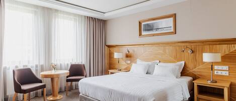 A hotel room with a large bed, two bedside tables, a round table with chairs, and a large window with curtains.