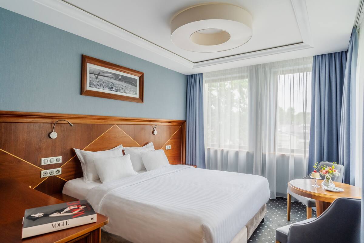 A hotel room with a large bed, a bedside table with a book, a small table with a vase, and a window with curtains.
