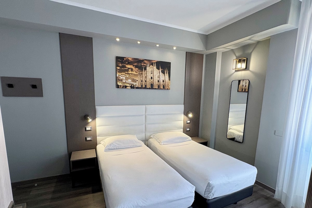 A hotel room with two single beds, a nightstand, a wall-mounted mirror, and a painting of a cityscape.