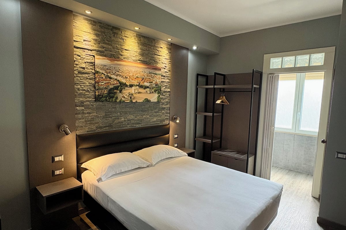 A modern bedroom with a large bed, a stone wall mural, a wooden wardrobe, and a view of the outdoors.