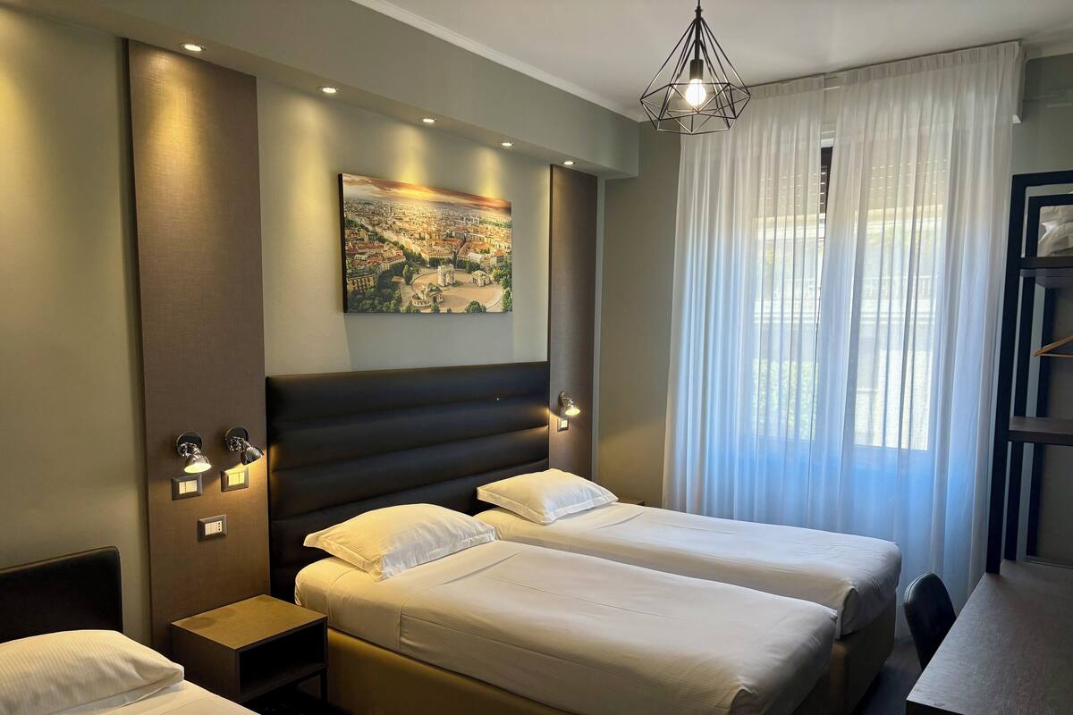 A hotel room with two beds, a desk, and a large window with sheer curtains.