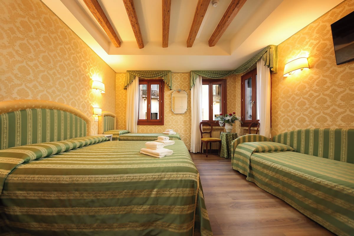 A hotel room with two single beds, a small table with a chair, a window with curtains, and a wall-mounted TV.