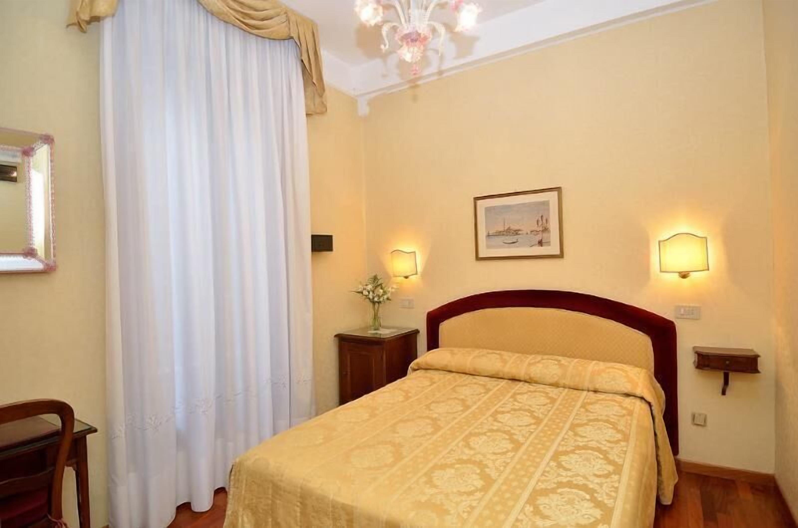 A hotel room with a bed, two bedside lamps, a nightstand with a vase, a framed picture on the wall, and a chandelier.