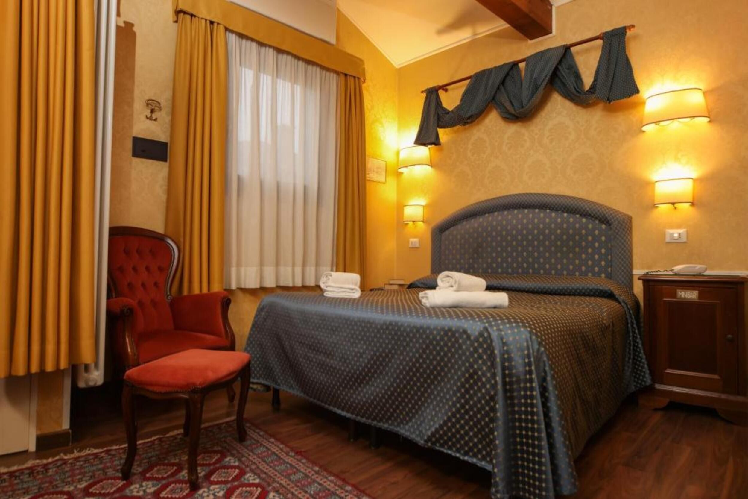 A hotel room with a bed, a chair, a nightstand, and two wall-mounted lamps.