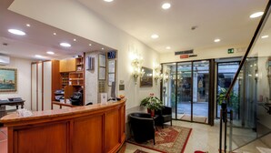 Zona de recepción de un hotel con un mostrador de madera, un reloj digital, una planta en maceta y una puerta de cristal que da a la entrada.