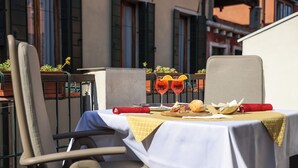 Un espacio de comedor al aire libre con una mesa dispuesta para dos personas, con mantel blanco, dos vasos de bebida roja, una hogaza de pan y un paño de papel rojo.