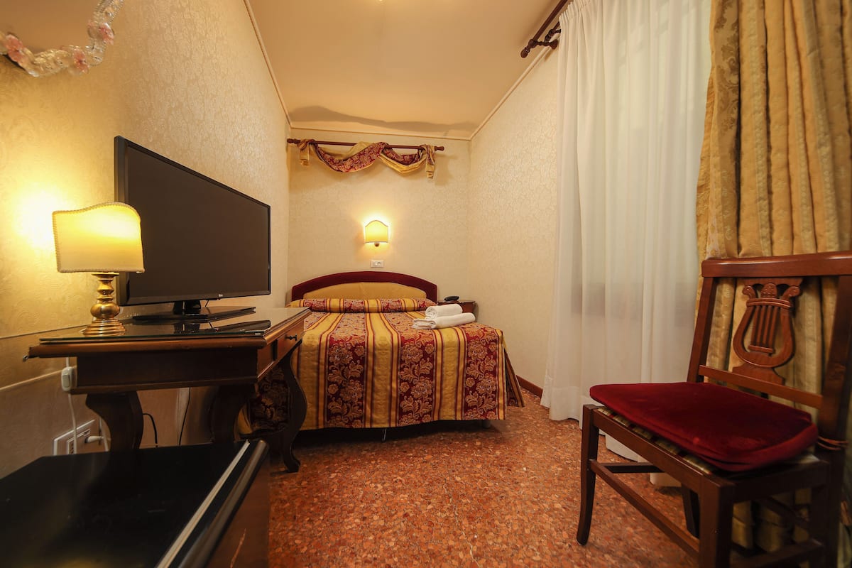 A hotel room with a bed, a desk with a lamp, a television, a chair, and a wall-mounted telephone.