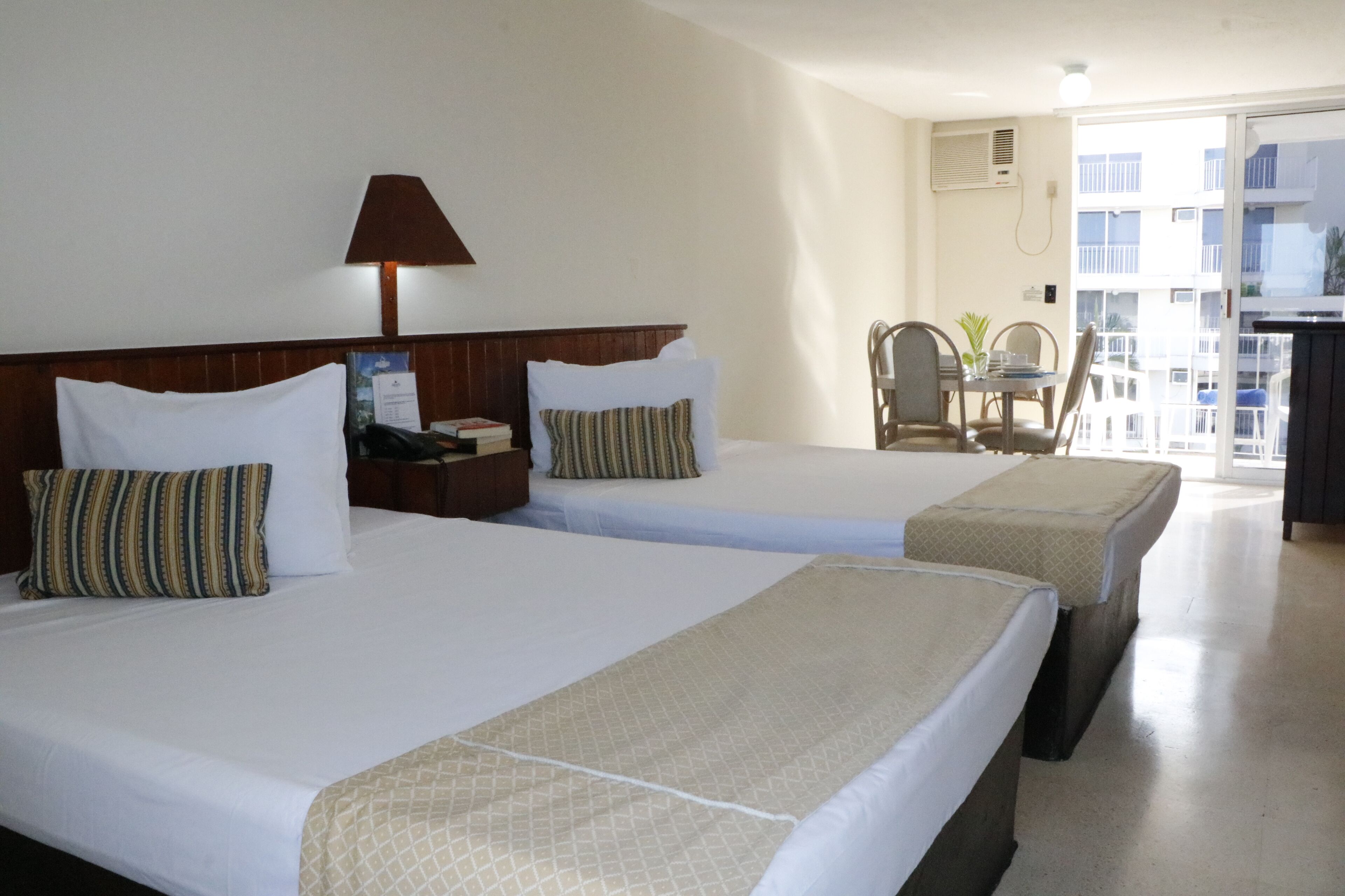 A hotel room with two beds, a desk, a chair, and a balcony with a view of the city.