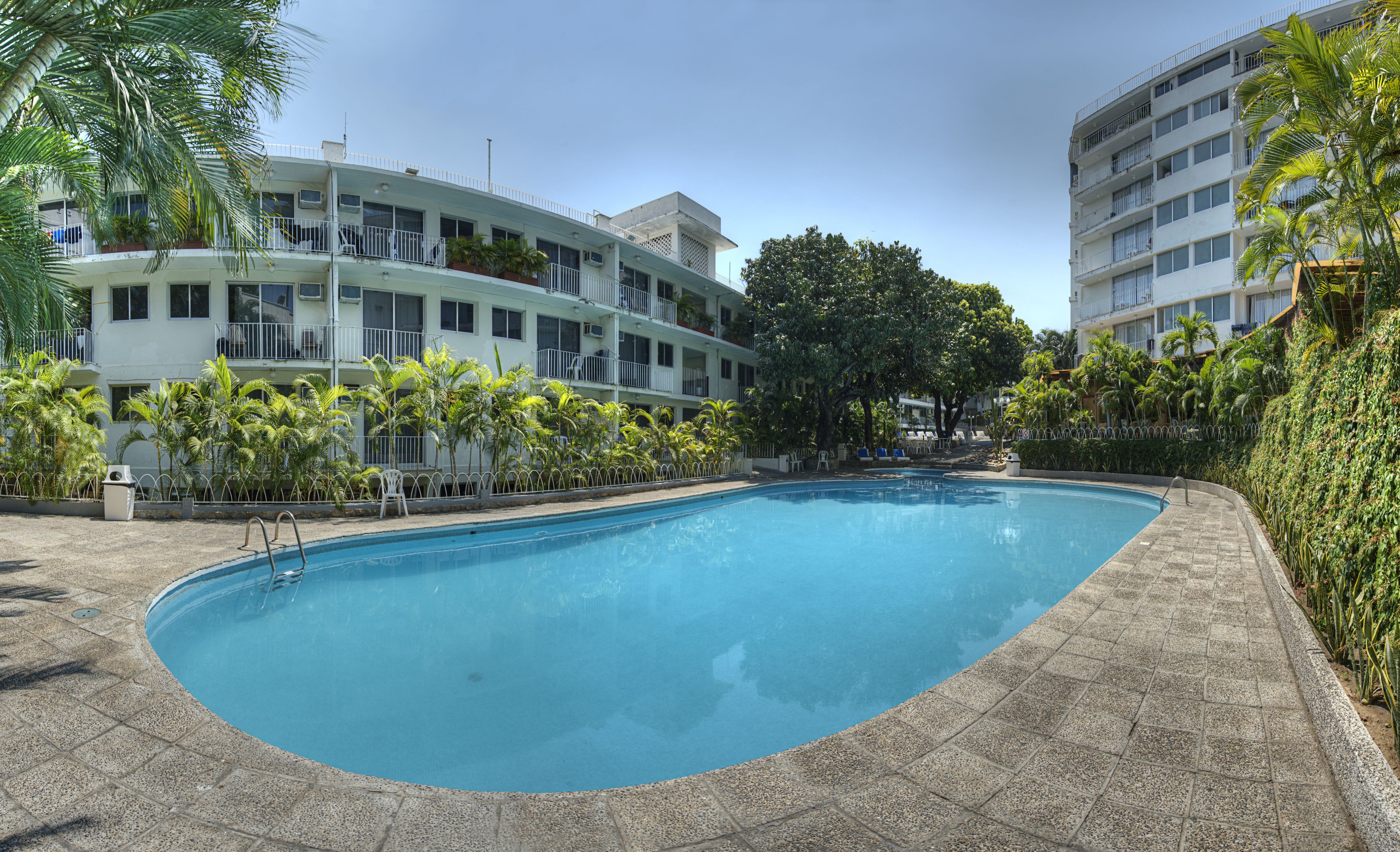 Una piscina con solárium de baldosas, rodeada de palmeras y un edificio de varios pisos con balcones.