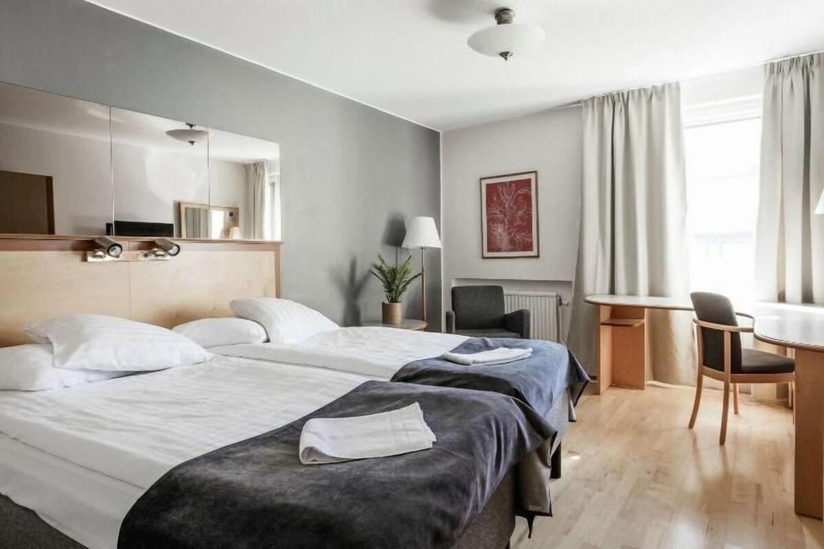 A hotel room with two beds, a desk, a chair, a lamp, and a window with curtains.