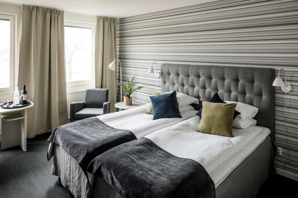 A hotel room with a large bed, two bedside tables, a chair, a potted plant, and a window with curtains.