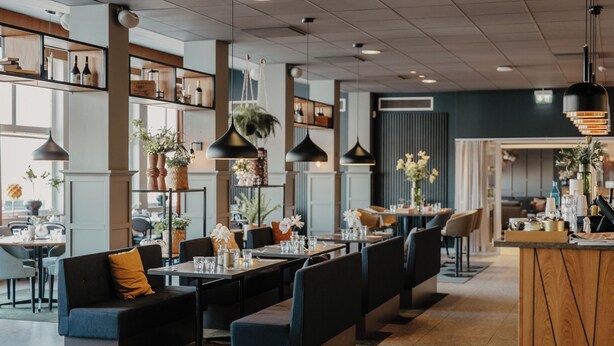 A modern restaurant interior with high tables, booths, hanging pendant lights, and potted plants.