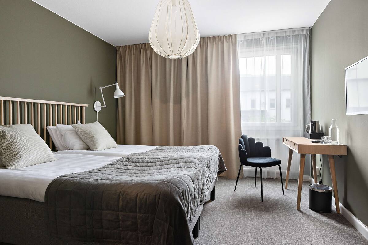 A hotel room with a bed, bedside table, lamp, chair, and a view of the outside through a window.