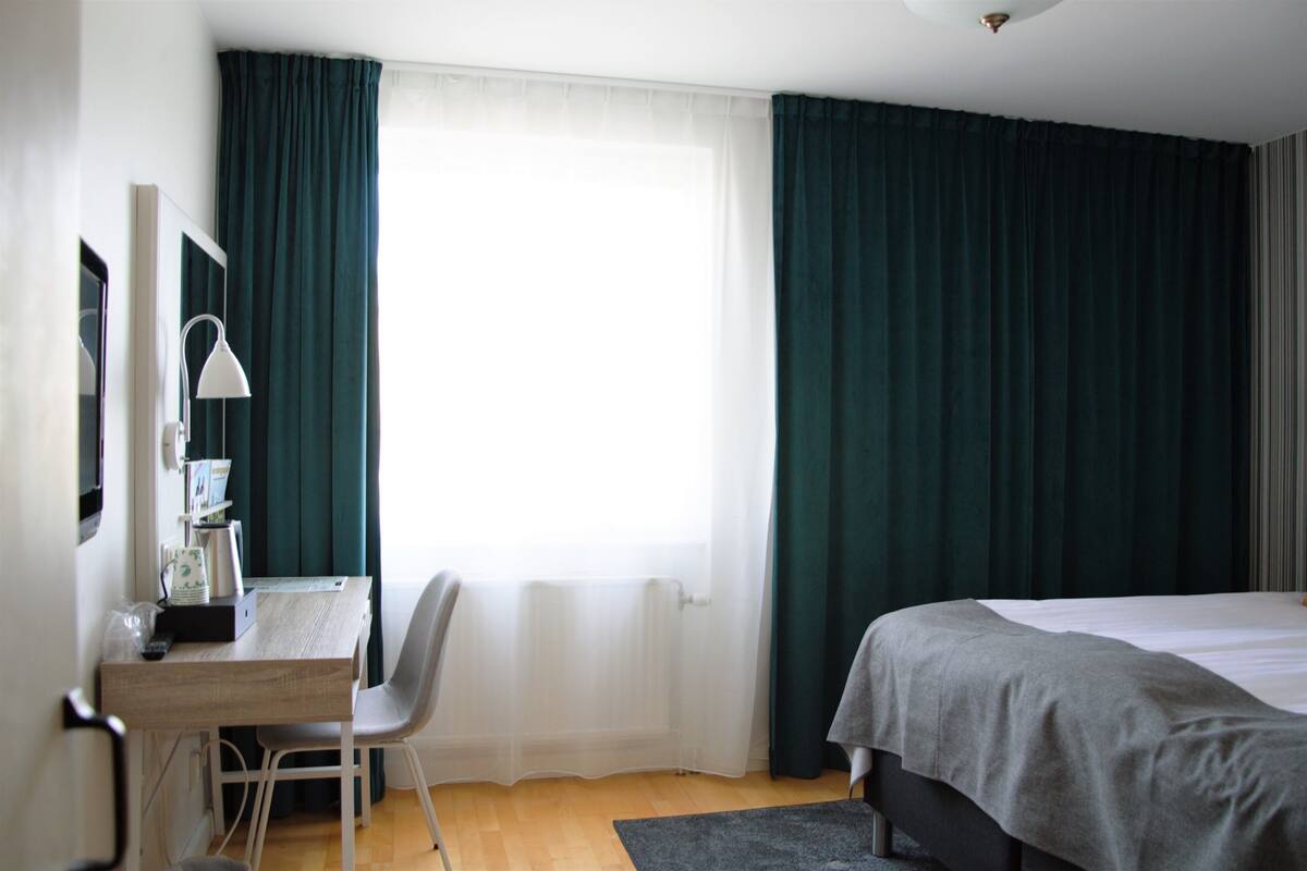 A hotel room with a bed, a desk with a chair, a lamp, and a window with curtains.