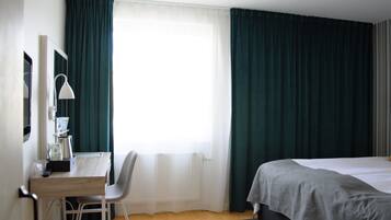 Une chambre d’hôtel avec un lit, un bureau avec une chaise, une lampe et une fenêtre avec des rideaux.