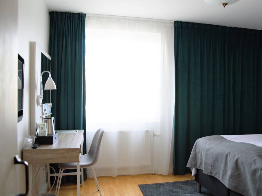 Une chambre d’hôtel avec un lit, un bureau avec une chaise, une lampe et une fenêtre avec des rideaux.