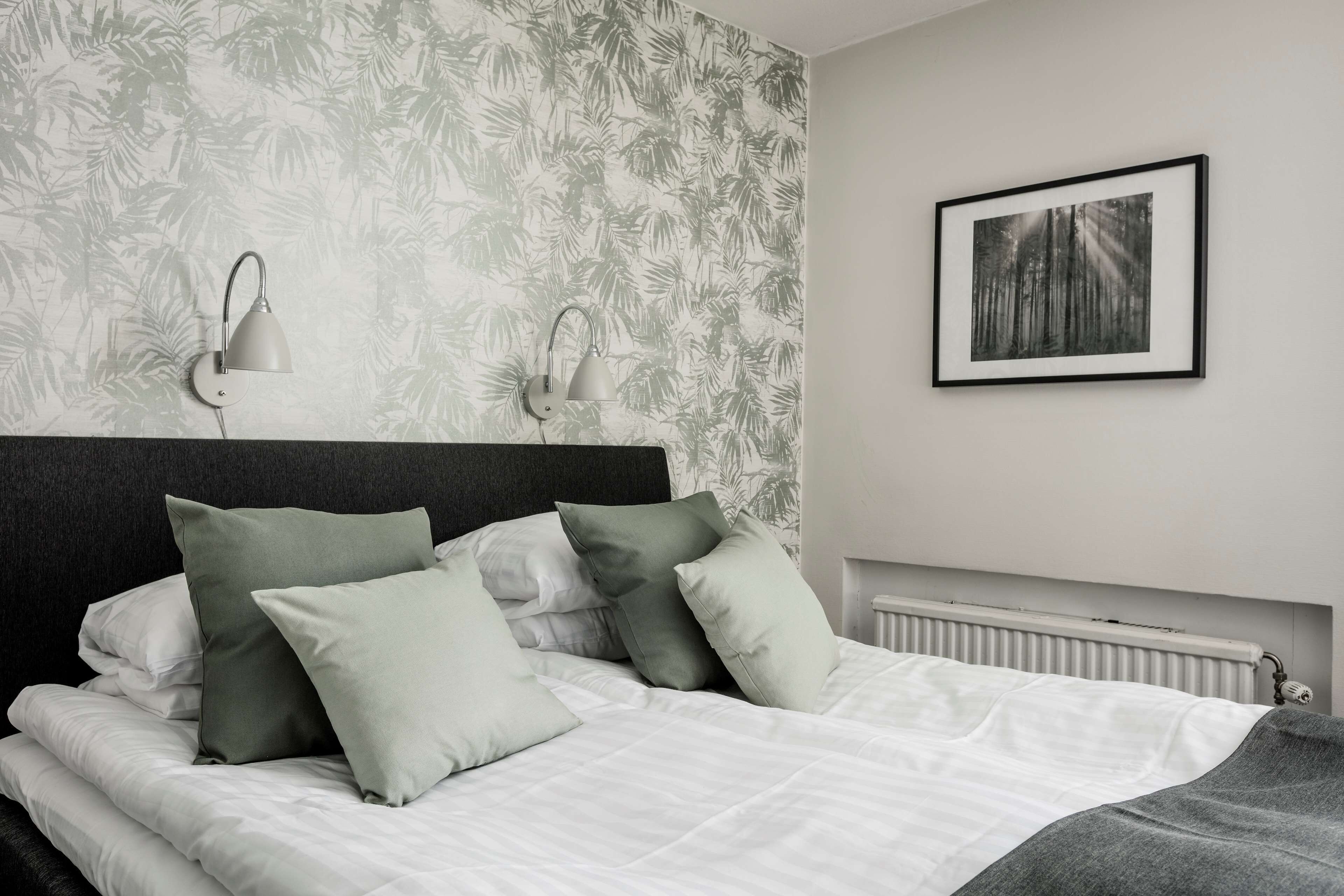 A neatly made bed with white linens and green pillows, a headboard, and a wall with a tropical leaf pattern. There are two wall-mounted lamps and a framed picture of a forest.