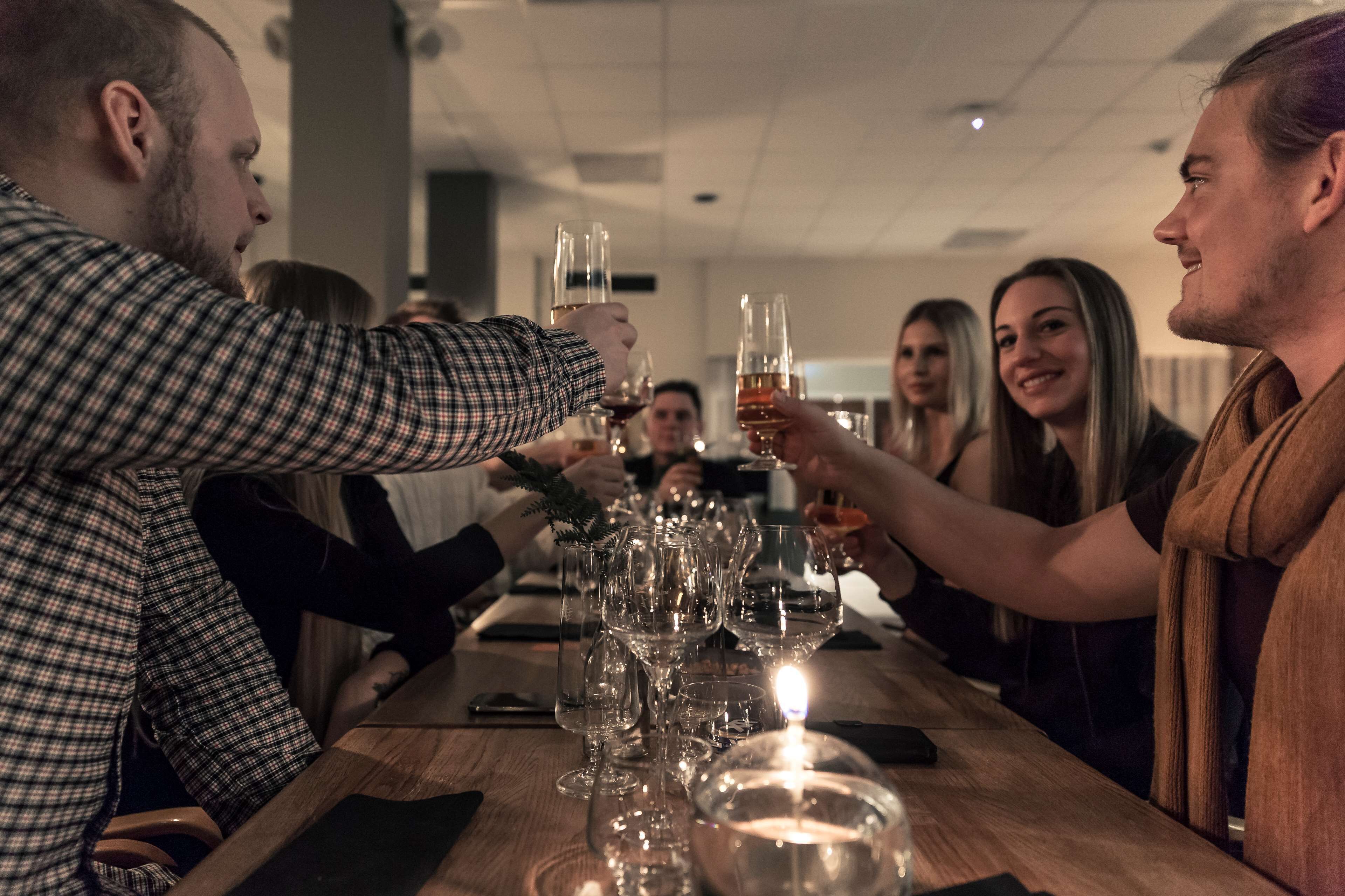 A group of people toasting at a wooden table with wine glasses and a candle, in a room with a high ceiling and visible columns.