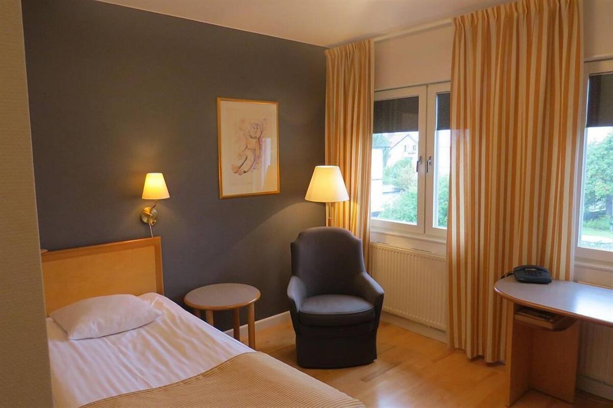 A hotel room with a bed, a chair, a desk, a lamp, a picture on the wall, and a window with curtains.