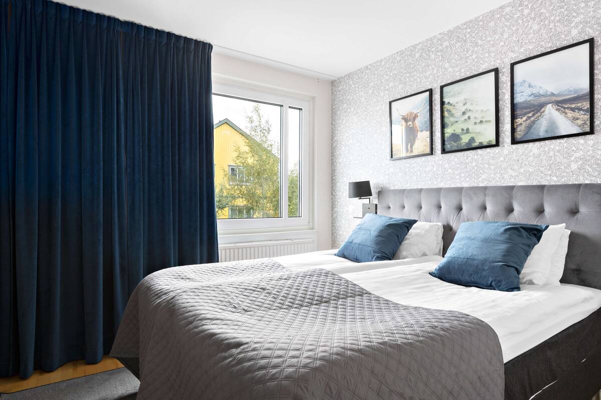 A bedroom with a large bed, a window with curtains, a wall with framed pictures, and a bedside table with a lamp.