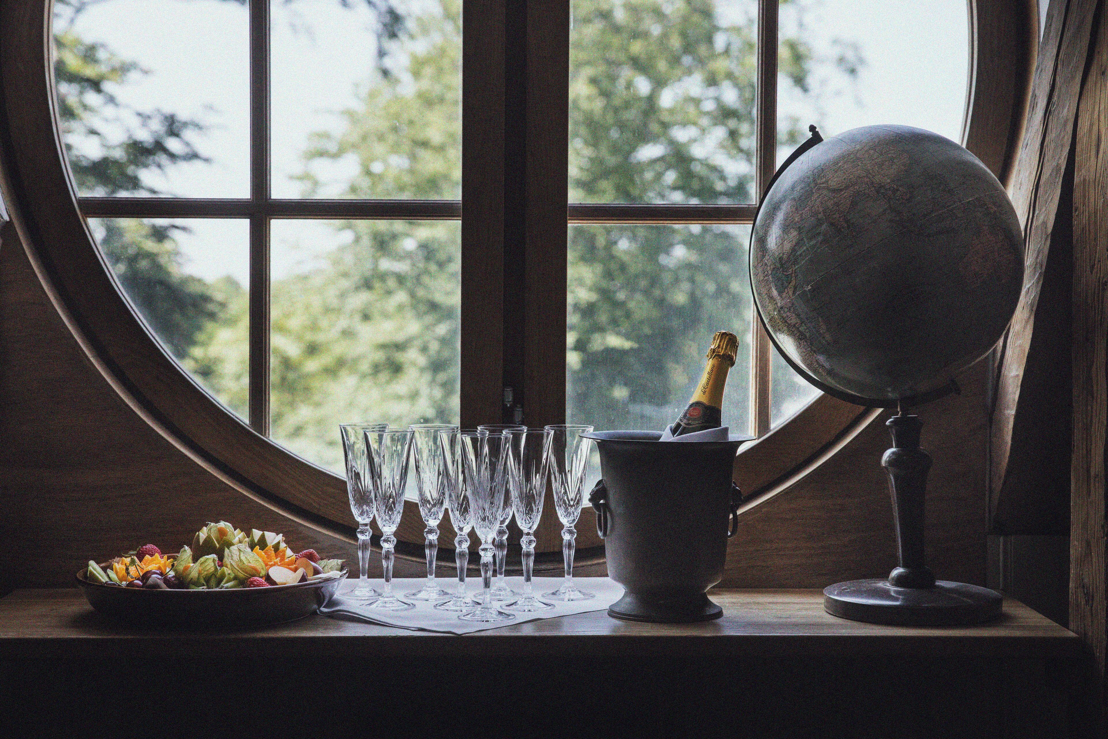 A table with a globe, champagne bottle, glasses, and a fruit bowl by a window with a view of trees.