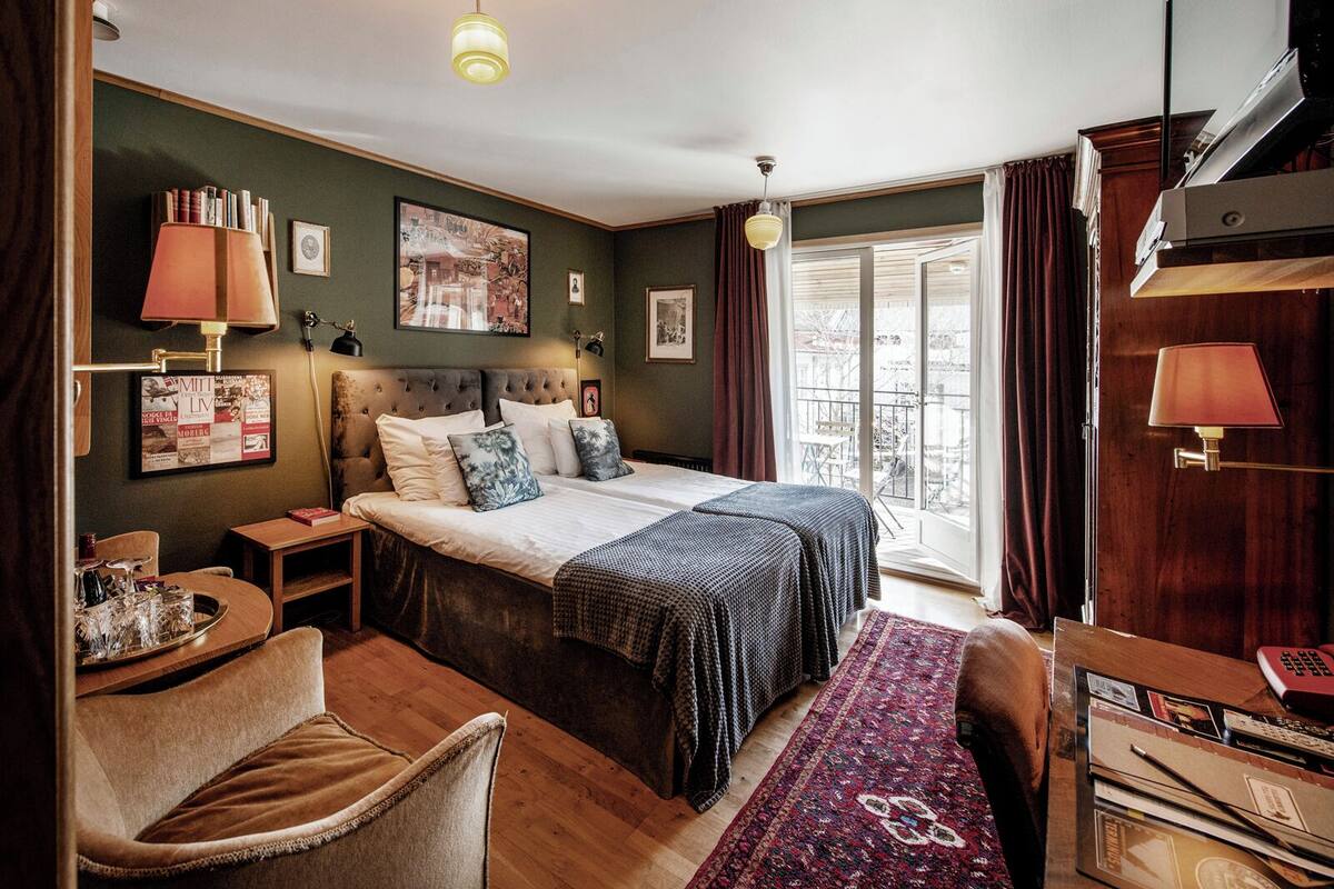 Ein helles Schlafzimmer mit einem großen Bett, einem Schreibtisch und einem Stuhl. Ein Fenster mit Vorhängen, ein Bücherregal und ein gerahmtes Bild sind an der Wand zu sehen.